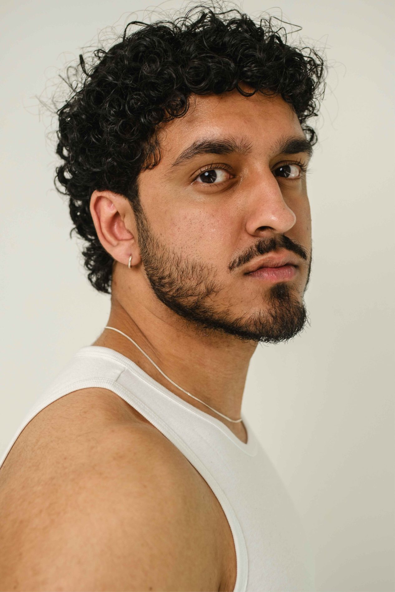 A chest up headshot on a cream background. They are about 20 with short curly brown hair, a short beard and moustache, and brown eyes. They are wearing a white vest, silver, chain, and a small silver hoop earring.
