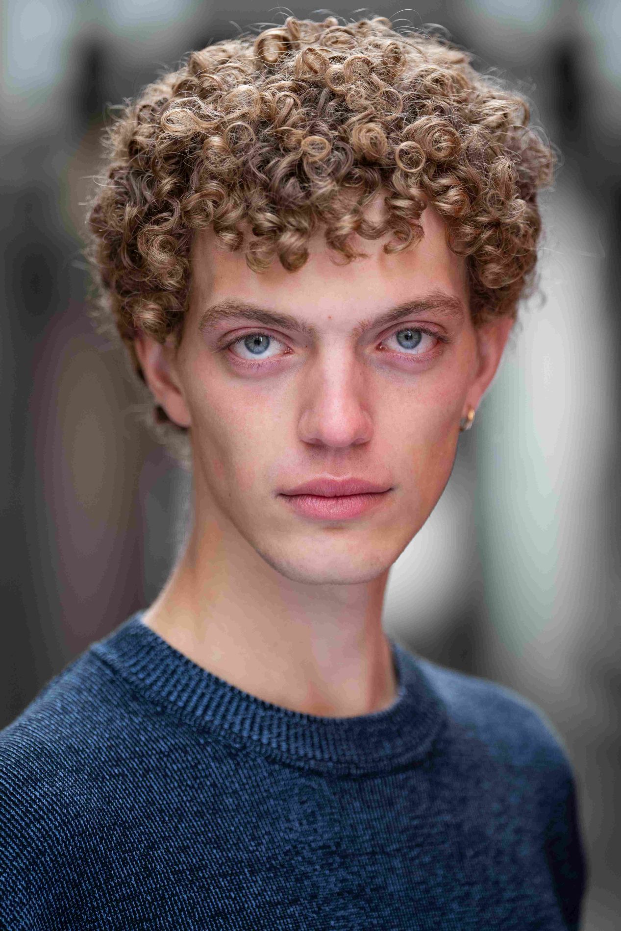A neck up headshot on a blurred background. They are about 20 with short curly dirty blonde hair and blue eyes. They are wearing a dark blue jumper and a small gold hoop earring.