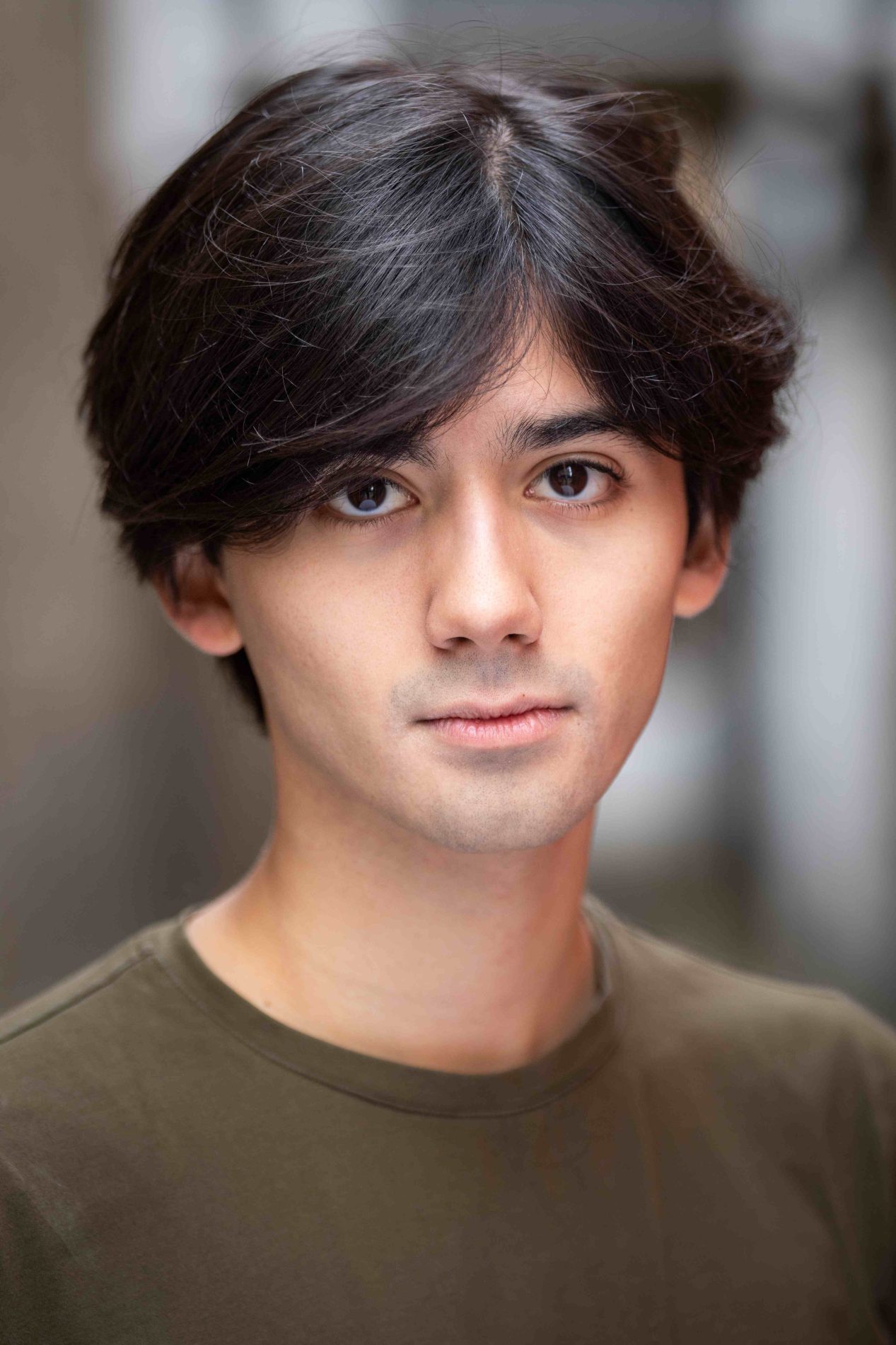 A neck up headshot on a blurred background. They are about 20 with short dark brown hair and brown eyes. They are wearing a brown shirt.