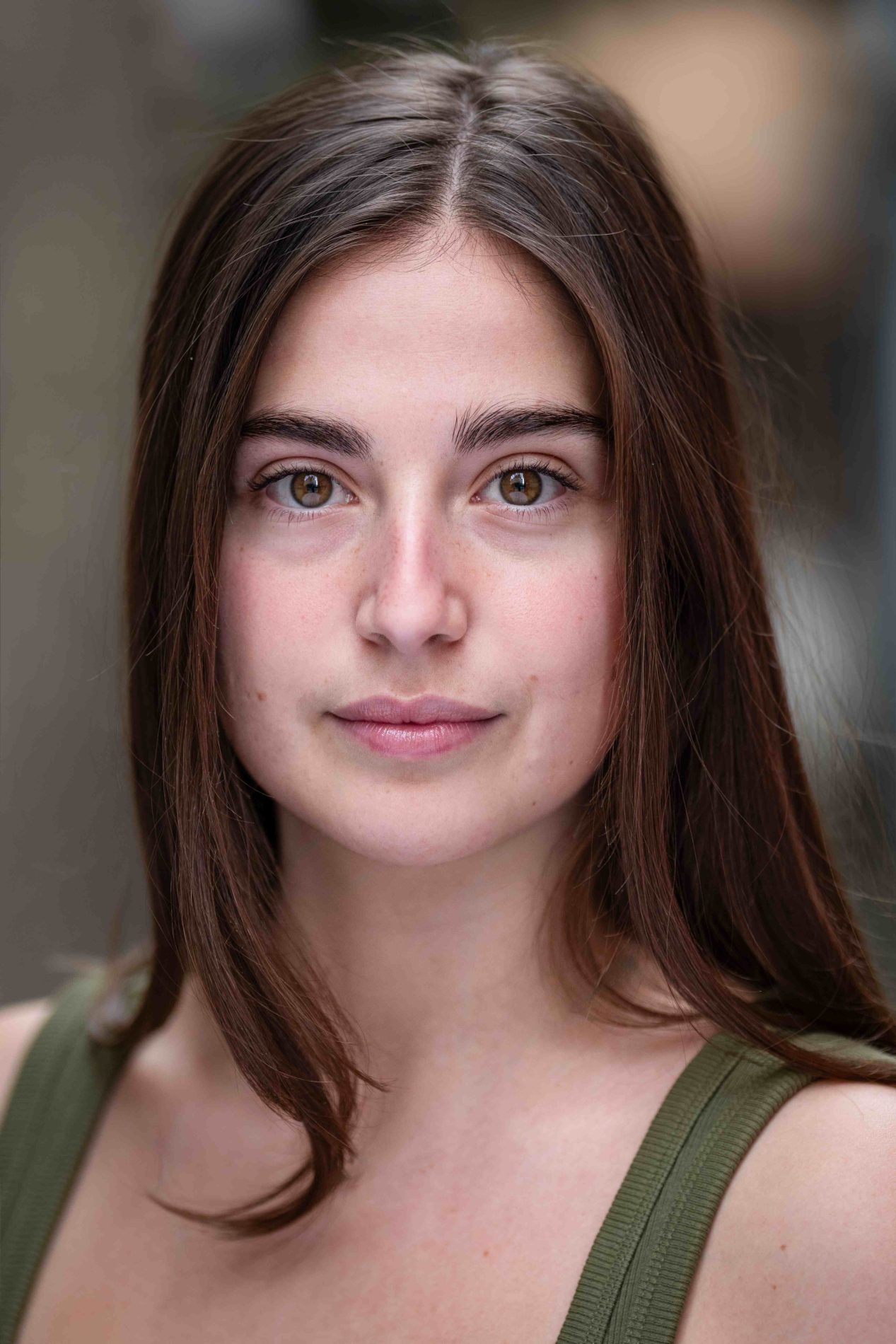 A neck up headshot on a blurred background. They are about 20 with long brown hair and brown eyes. They are wearing a green camisole.