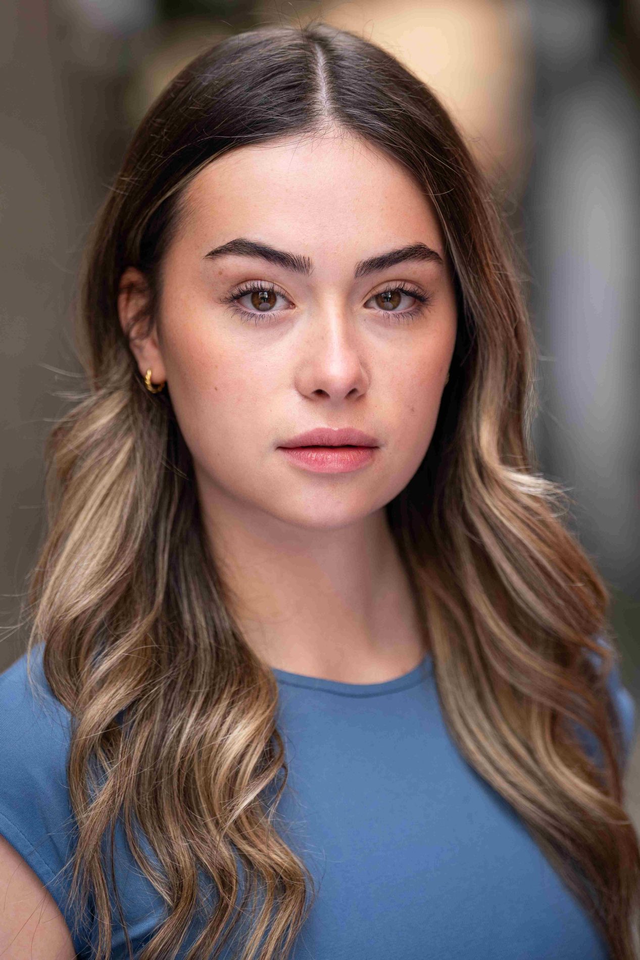 A neck up headshot on a blurred background. They are about 20 with long brown hair and brown eyes. They are wearing a blue top and small gold hoop earrings.
