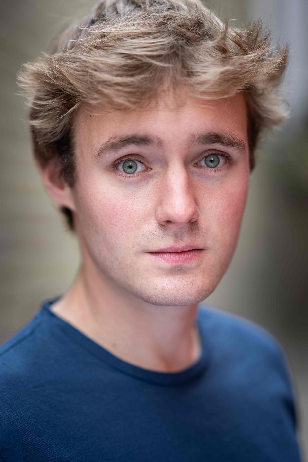A neck up headshot on a blurred background. They are about 20 with short blonde hair and blue eyes. They are wearing a blue t-shirt.