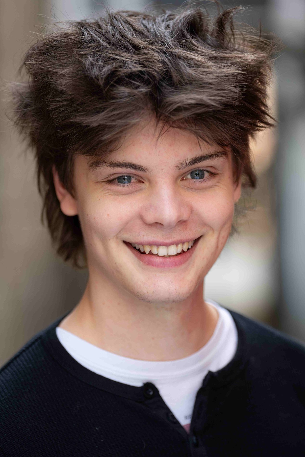 A neck up headshot on a blurred background. They are about 20 with short brown hair and blue eyes. They are wearing a black jumper with a white shirt underneath.