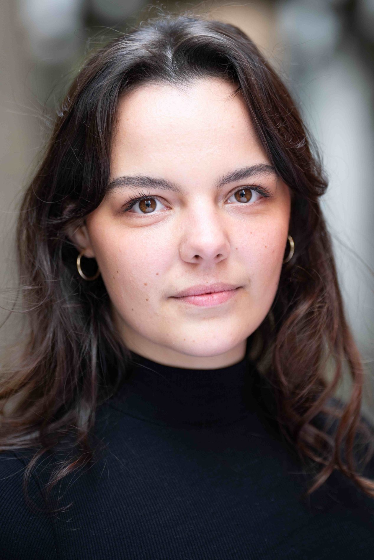 A neck up headshot on a blurred background. They are about 20 with medium length brown hair and brown eyes. they are wearing a black top and gold hoop earrings.