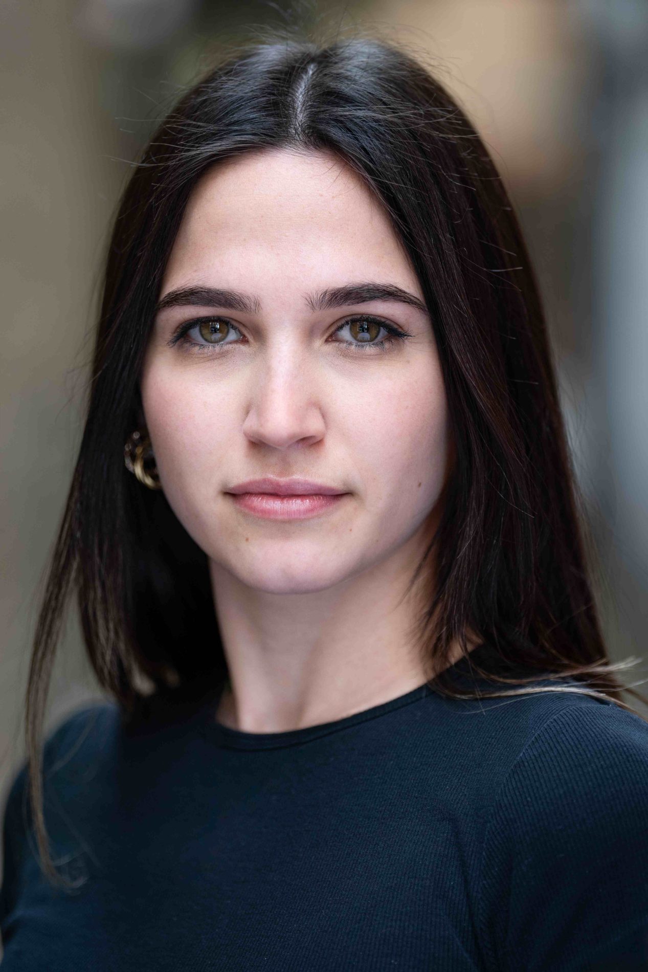 A neck up headshot on a blurred background. They are about 20 with long dark brown hair and hazel eyes. They are wearing a black top and gold earrings.