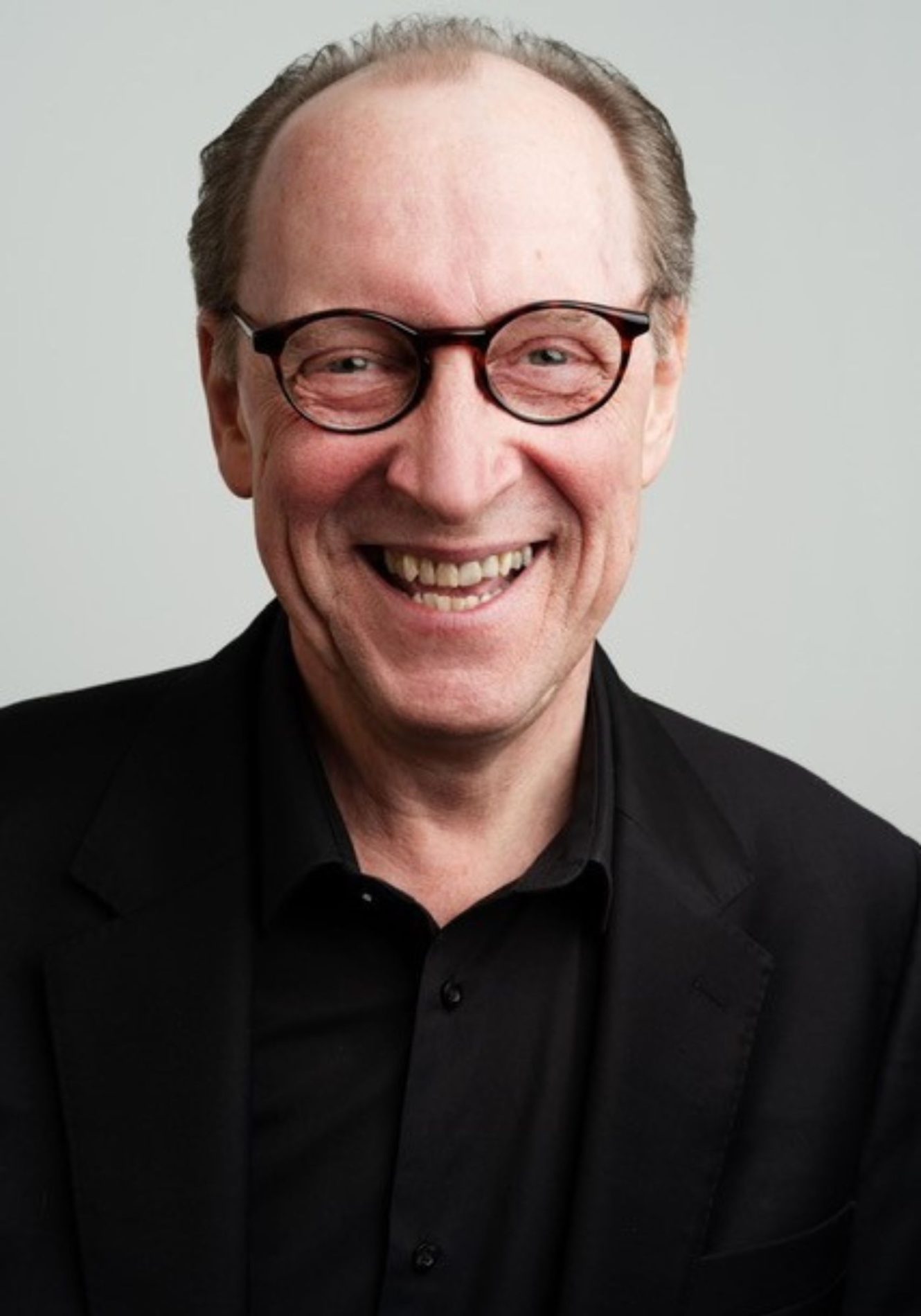 A chest up headshot on a light grey background. They are middle aged with short grey hair, brown eyes, and tortoise shell framed glasses. They are wearing a black button down shirt with a black suit jacket.