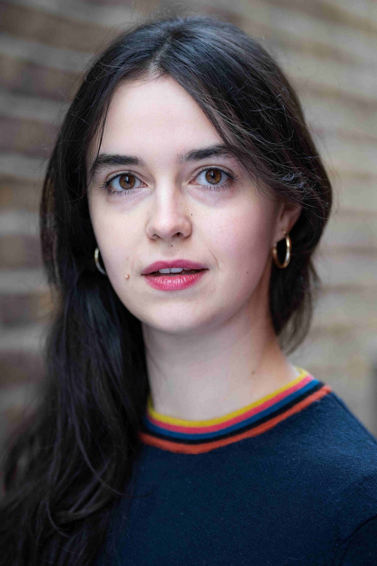 A neck up headshot on a blurred background. They are about 20 with long dark brown hair and brown eyes. They are wearing a black jumper with a colourful collar and gold hoop earrings.