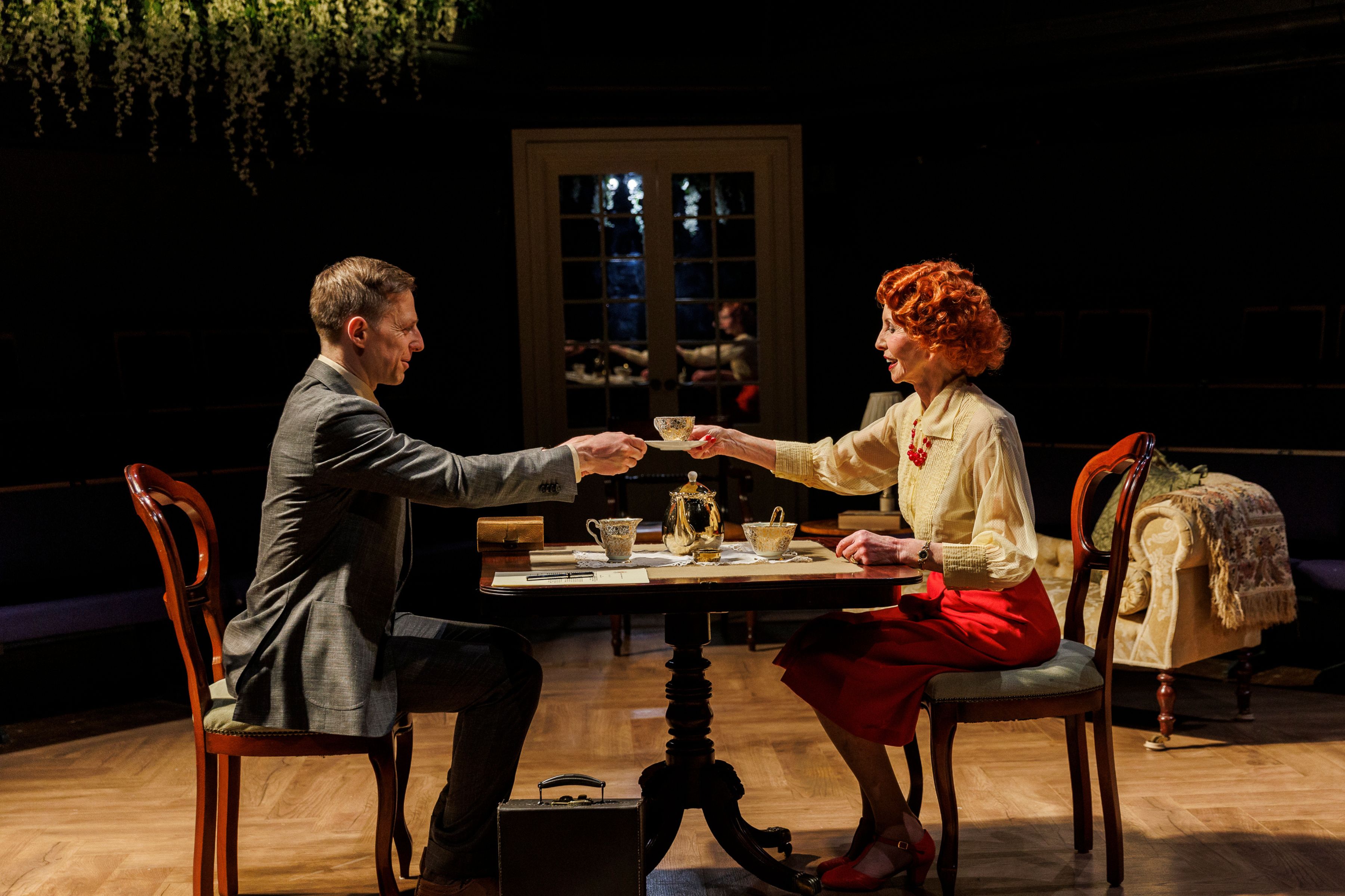 A man and woman are sat across from each other at a table. The man is wearing a grey suit and the woman is wearing a white blouse and red skirt. The woman is passing the man a teacup over the table.
