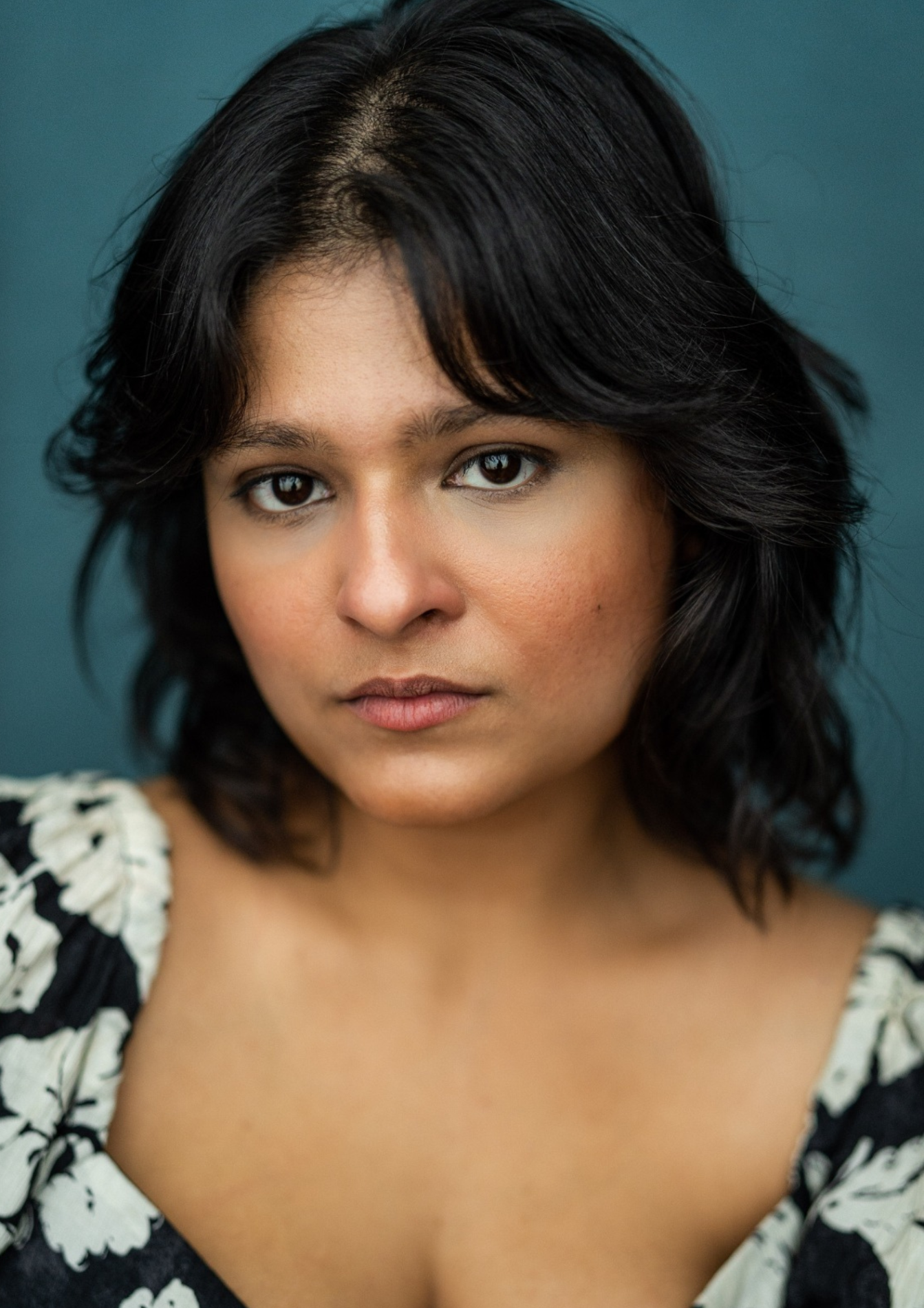 A headshot of a woman. She has tan skin and shoulder length wavy black hair and dark eyes. She wears a black and white floral blouse.