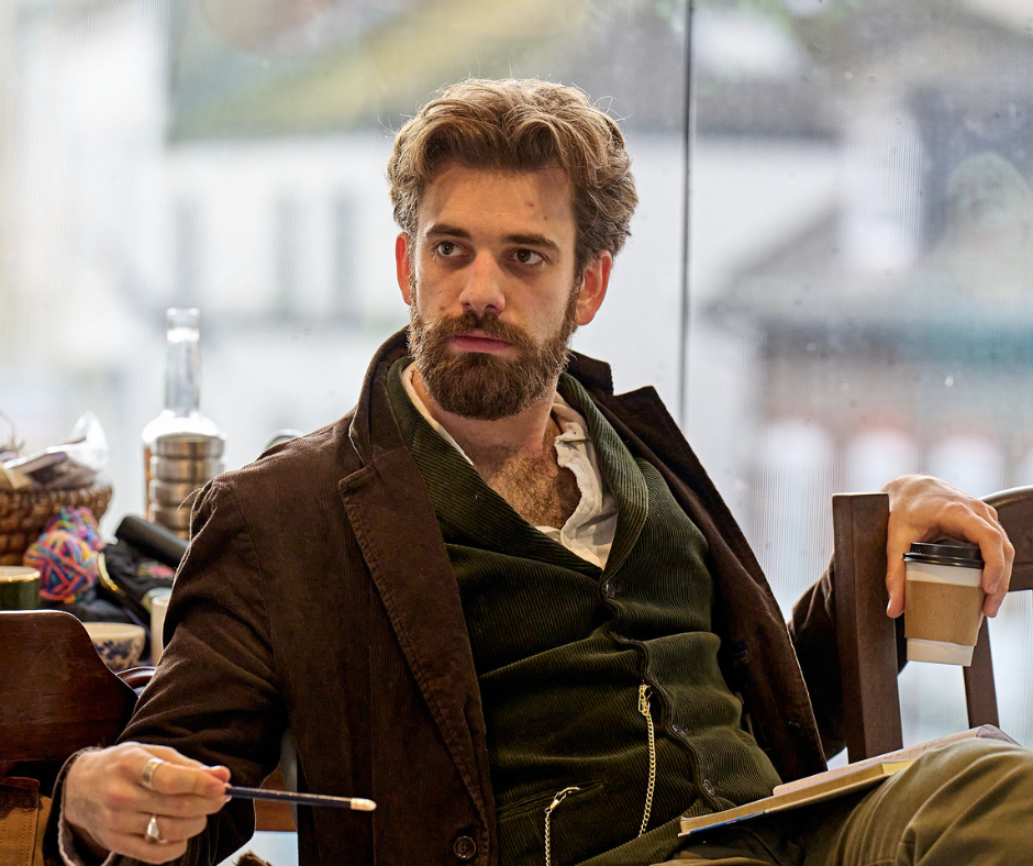Andrew Richardson sits by a window wearing a brown jacket and vest. He holds a to-go coffee cup in his hand and has an open book in his lap.