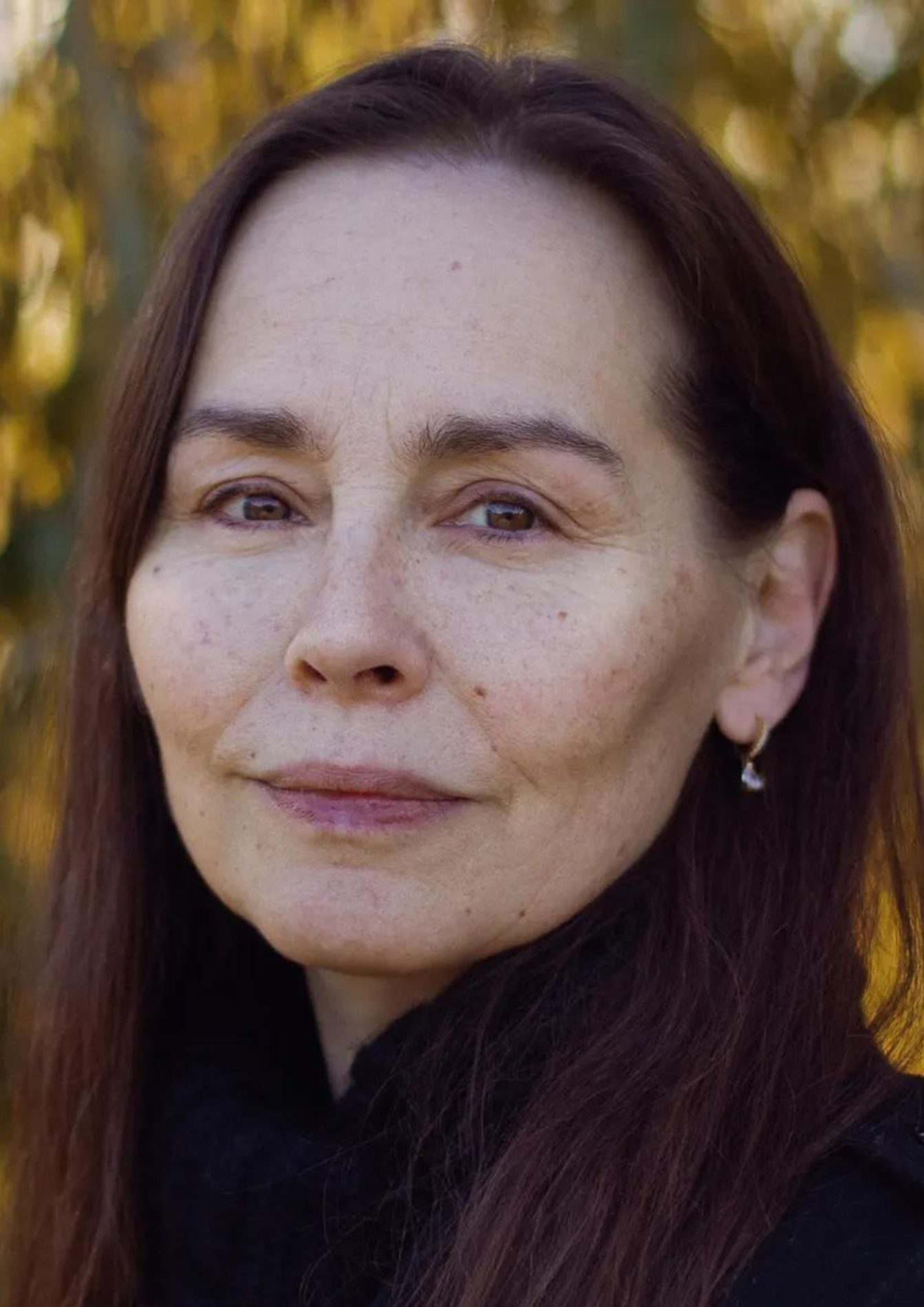 A woman with dark brown hair and brown eyes. She is wearing a black top and has gold hoop earrings in.