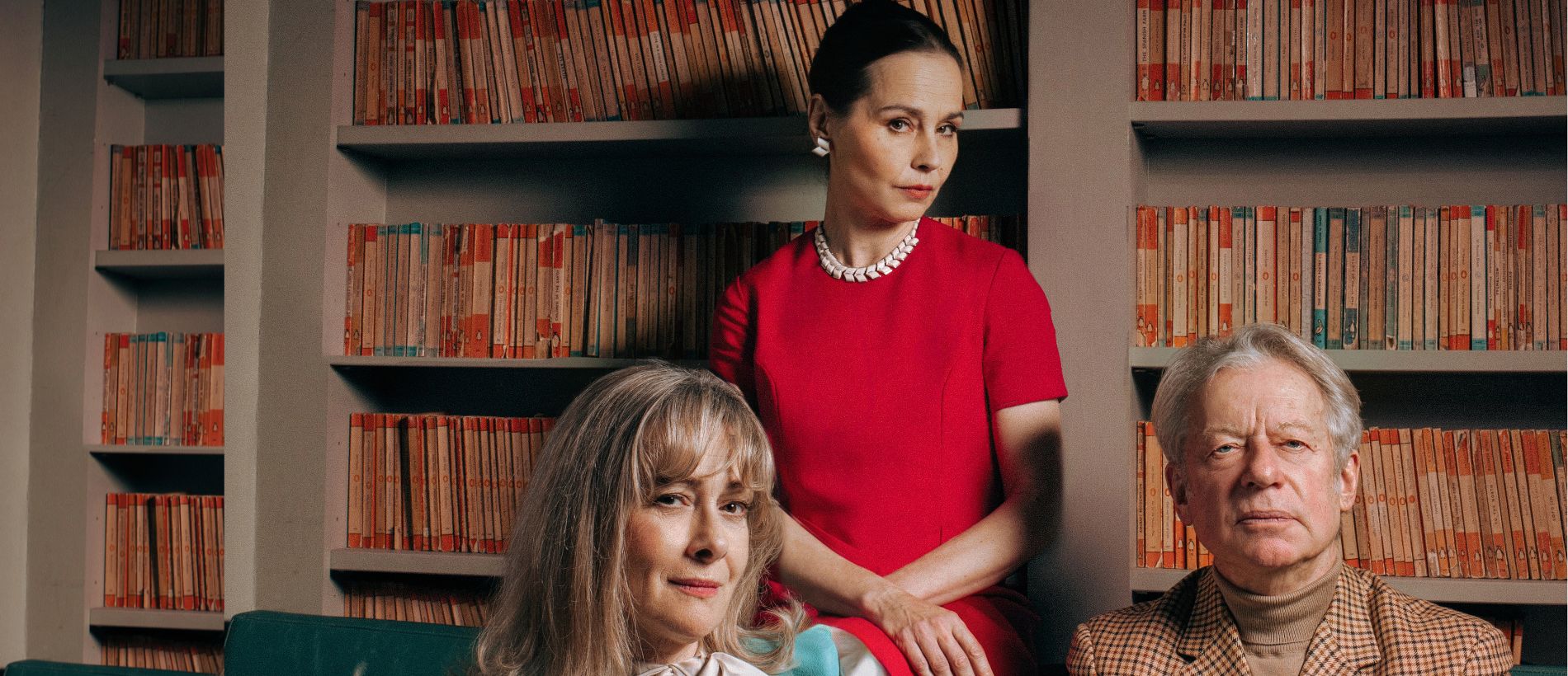 Two women and a man are in shot. The woman sat front left is wearing a blue trouser suit with a silk white shirt underneath. She has blonde hair to her shoulders with a fringe and is sat on a chair leaning on a table. The woman sat in the centre is wearing a pink dress with short sleeves and a pearly necklace. She is slightly higher than the other two people. She has dark brown hair which is styled in an updo. The man sat to the right is wearing a chequered neutral suit with a camel-coloured turtle neck. He has short grey hair and is staring directly to camera with his arms folded.