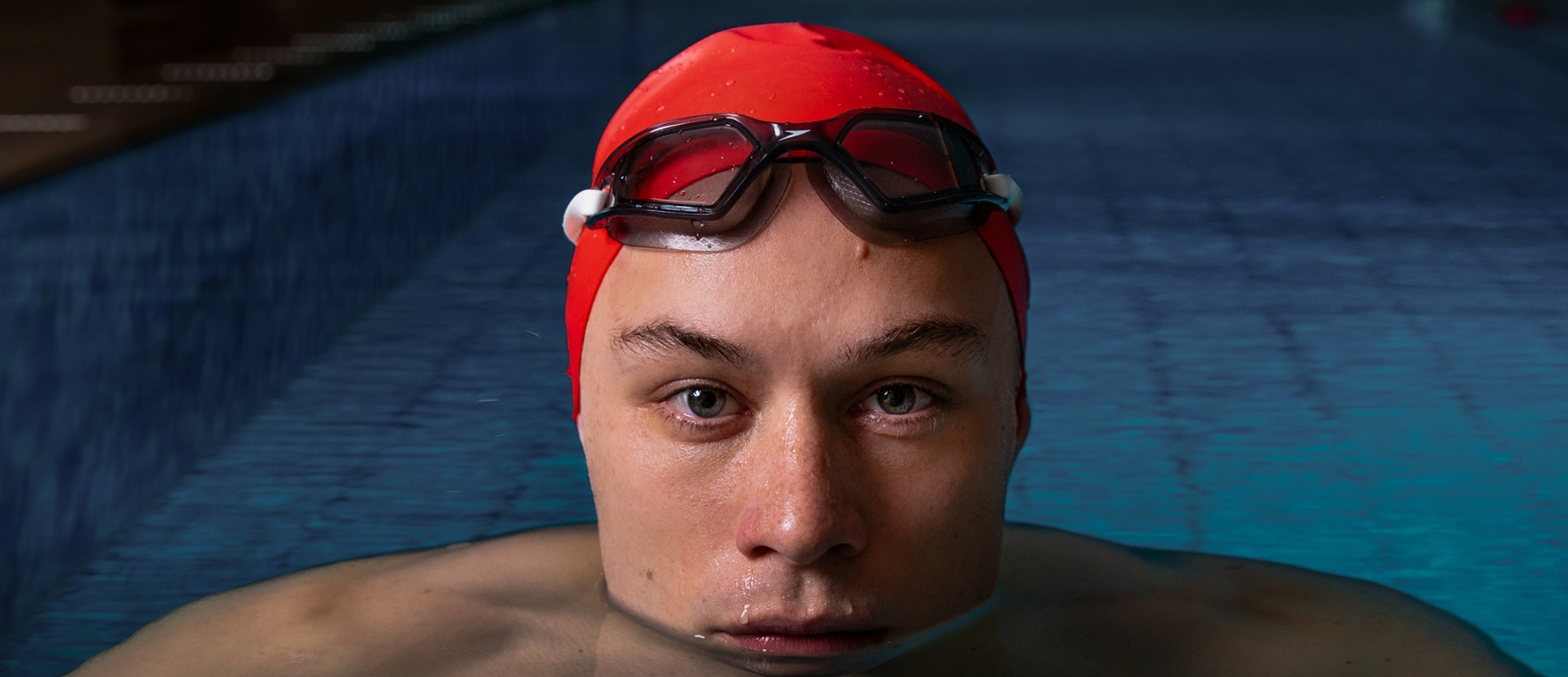 A man is seen in a swimming pool, submerged in water up to his chin. He wears a red swim cap with black swim goggles resting on his forehead. The length of the pool is visible behind him.
