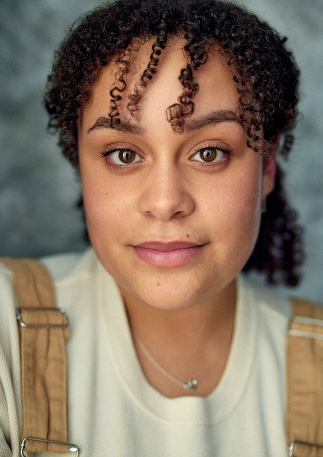 Portrait shot of a young woman with curly brown hair tied into a ponytail, brown eyes, and a pleasant expression on her face. She wears a cream coloured crew shirt with tan overall straps.
