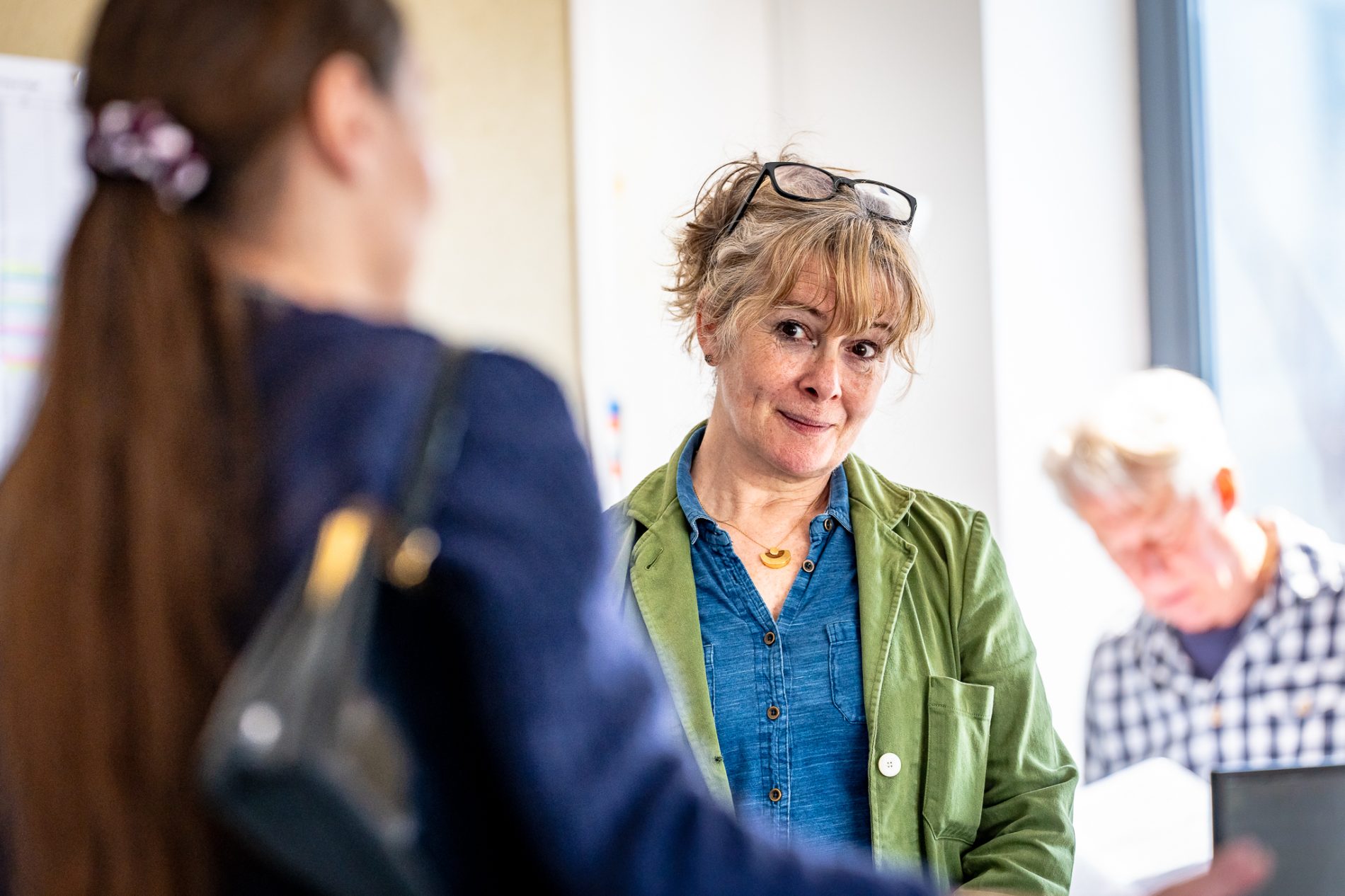 A mid-50s woman wearing a blue denim style shirt and green jacket stands in the middle with her head tilted looking thoughtfully off at something in the distance. She is framed by a woman on the left and man on the right who are out of focus.