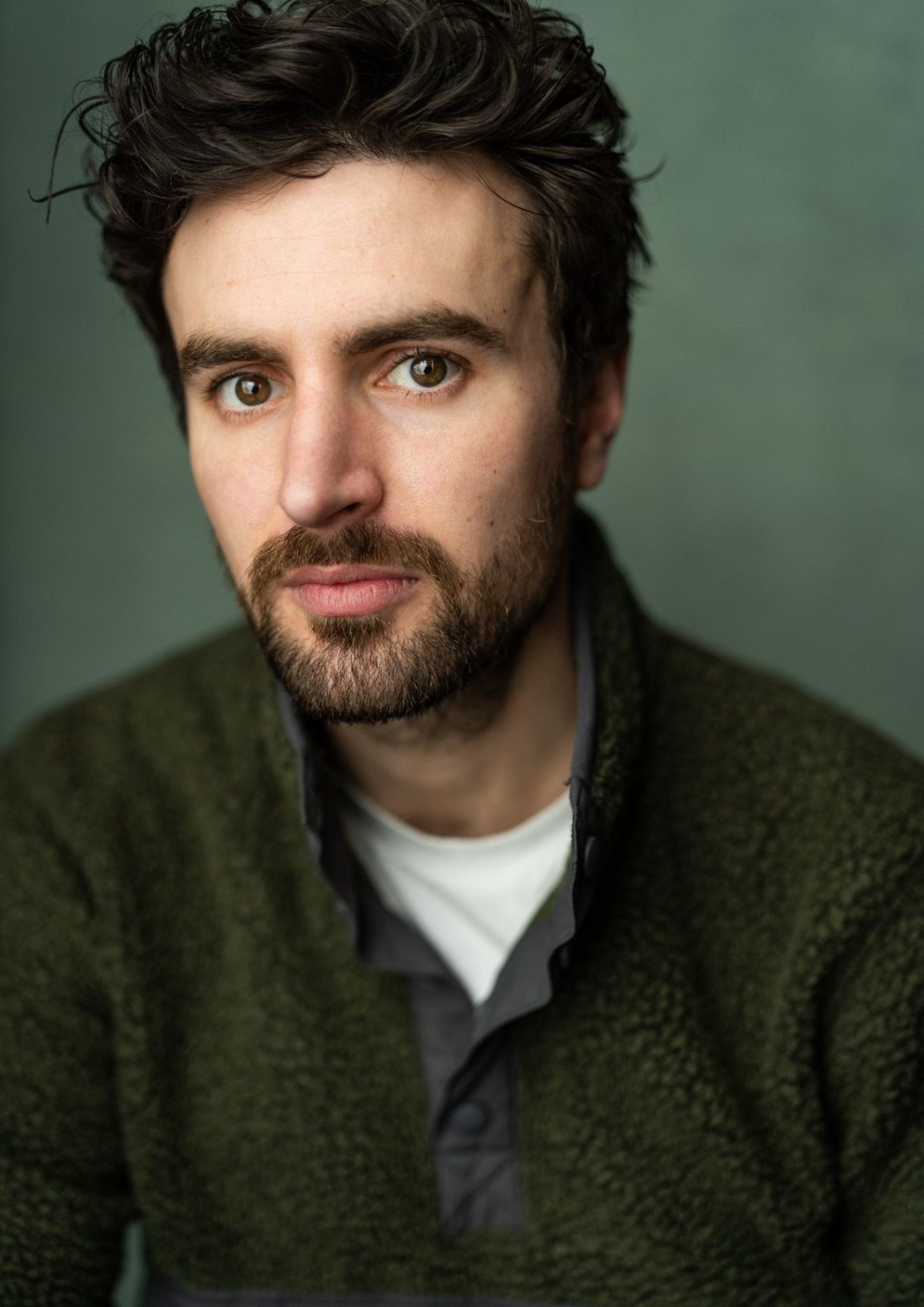 A chest up headshot on a green background. They are about 30 with short dark brown hair, hazel eyes, and a short beard and moustache. They are wearing a white t-shirt under a dark green jacket.