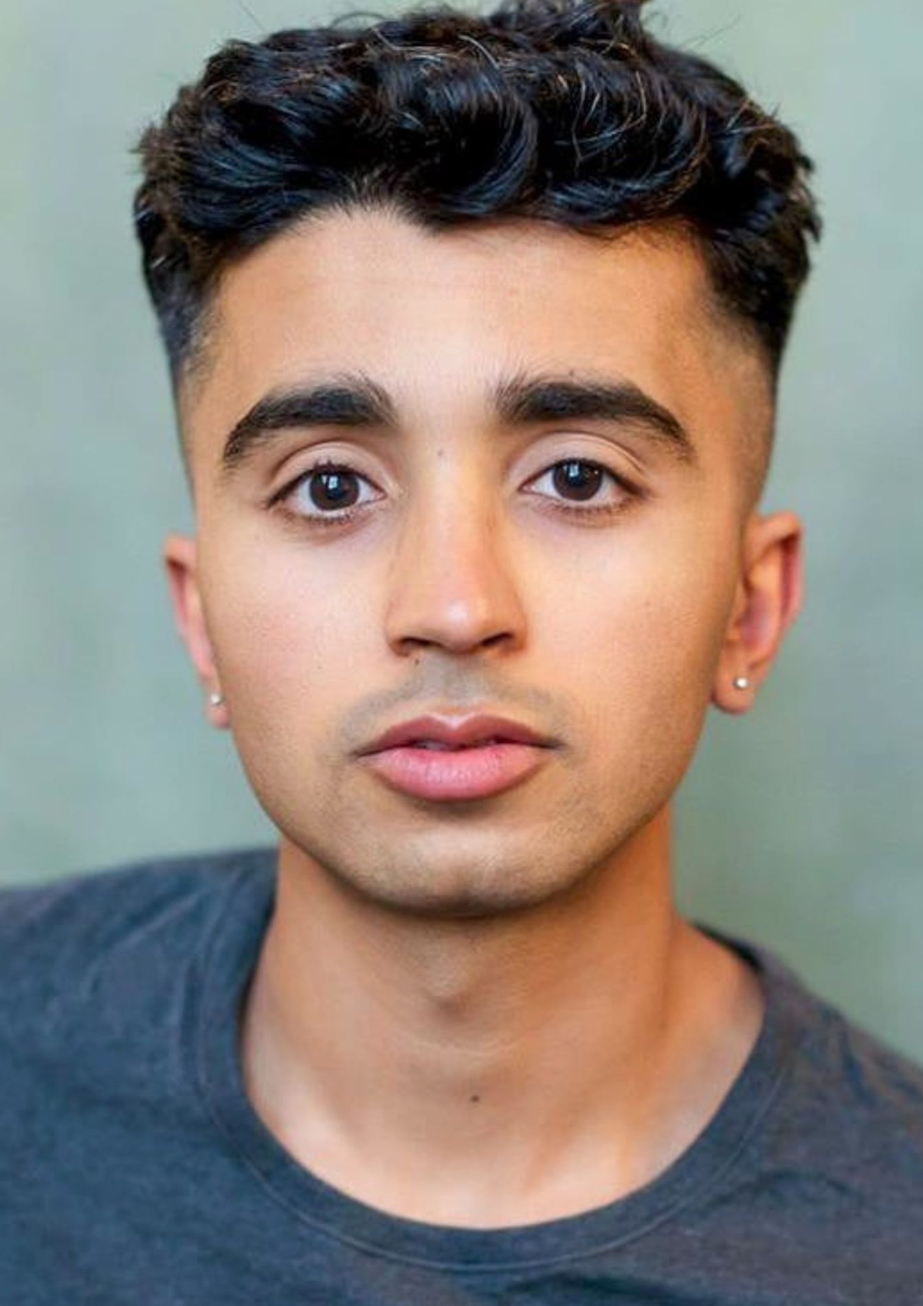 A neck up headshot on a blue-green background. They are about 25-30 with short dark brown hair, brown eyes, and a shadow of a moustache and beard. They are wearing a dark grey t-shirt and small diamond studs.