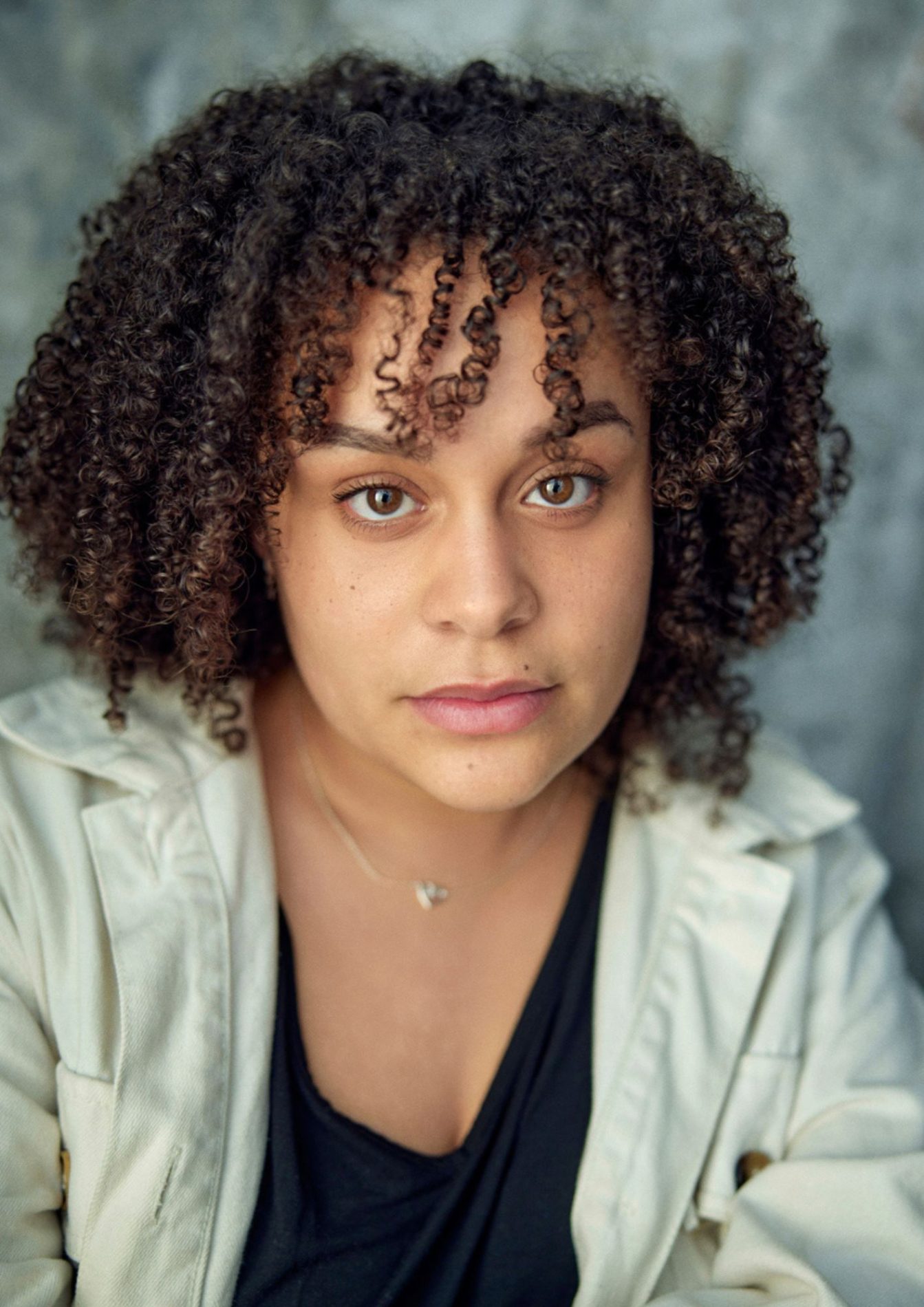 A chest up headshot of an actor on a grey background. They are about 30 with short brown curly hair and brown eyes. They are wearing a black shirt under a light green jacket with a silver necklace.