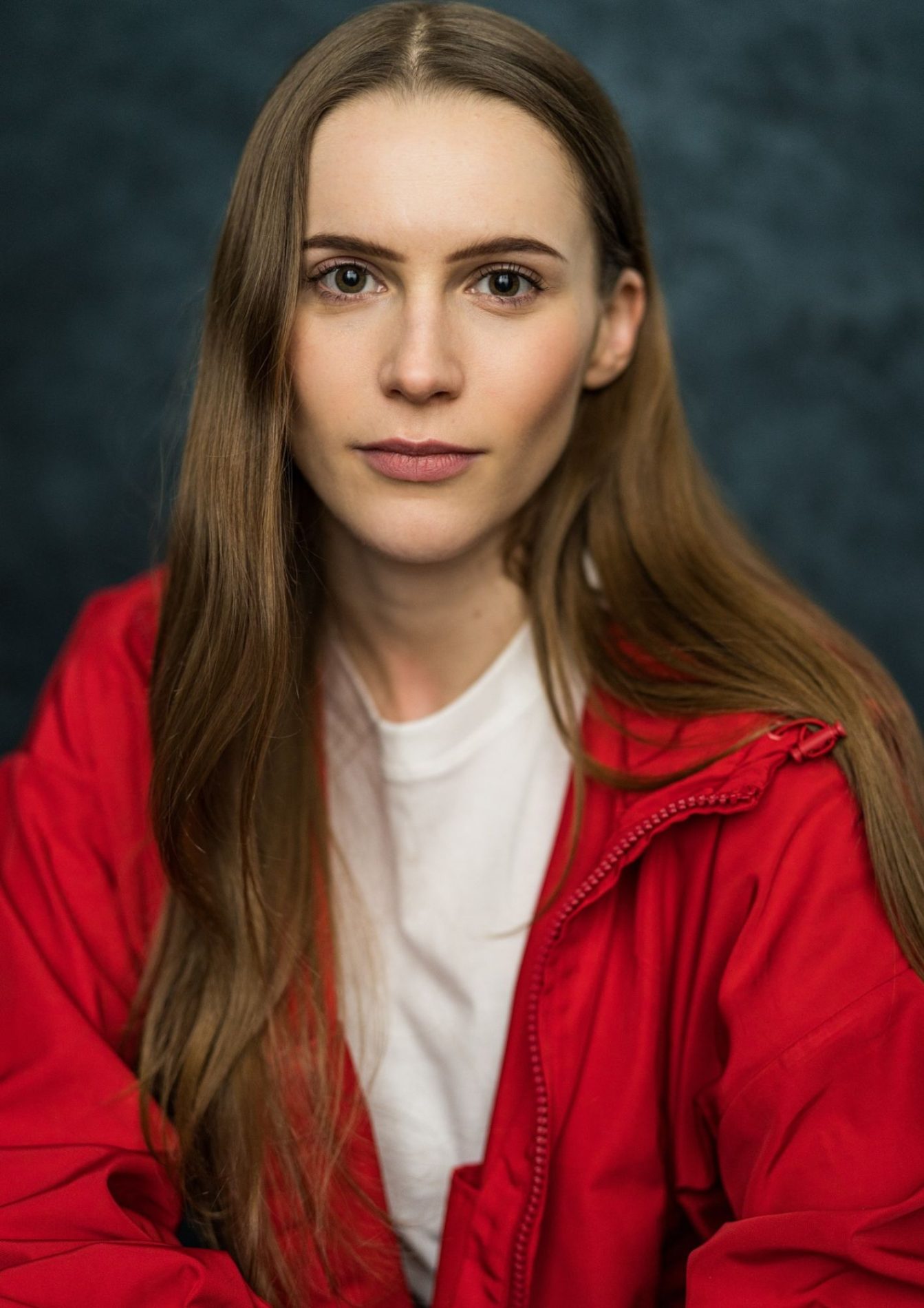 A chest up headshot on a dark grey background. They are about 25-30 with straight, long, brown hair and brown eyes. They are wearing a white t-shirt under a red zip up jacket.