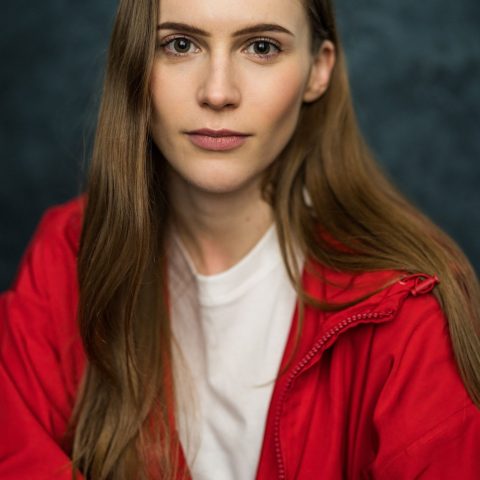 A chest up headshot on a dark grey background. They are about 25-30 with straight, long, brown hair and brown eyes. They are wearing a white t-shirt under a red zip up jacket.