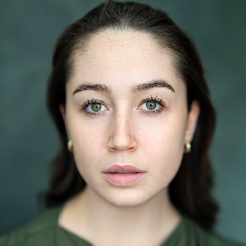 A neck up headshot on a dark green background. They are about 30 with dark brown hair and hazel eyes. They are wearing a green top and small gold hoops.