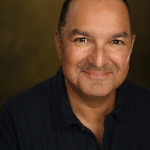 A chest up headshot on a brown background. They are middle aged with short brown hair and brown eyes. They are wearing a black collared shirt.
