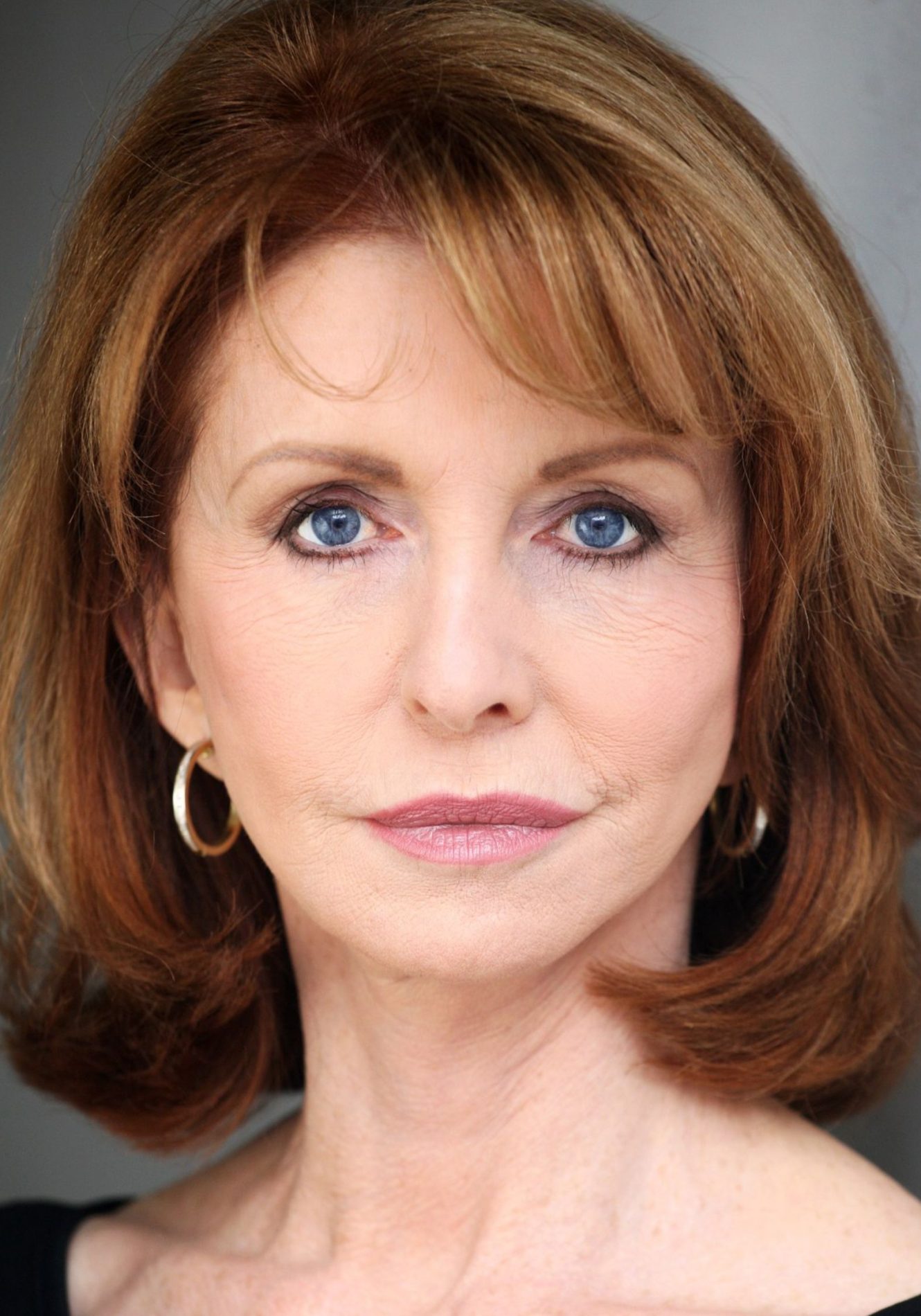 A neck up headshot on grey background. They are middle aged with shoulder length brown hair and blue eyes. They are wearing a black top and gold hoops with light glam makeup.
