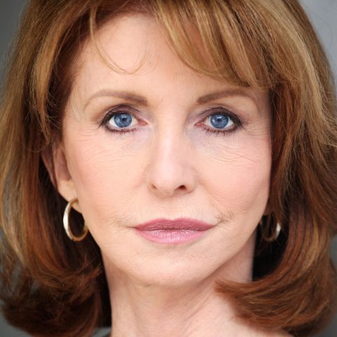 A neck up headshot on a grey background. They are older with shoulder length brown hair and blue eyes. They are wearing a black top and gold hoops with light glam makeup.