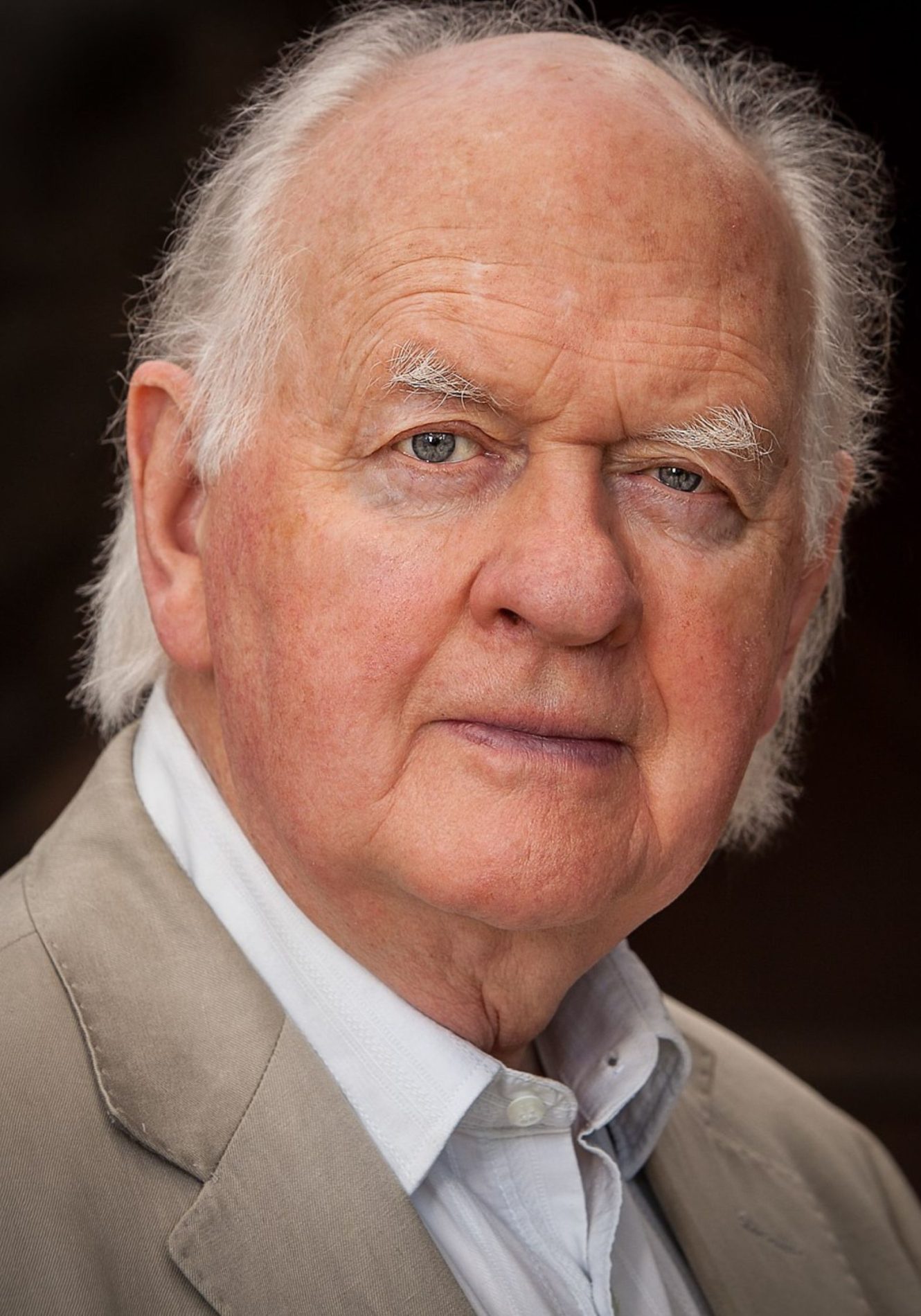 A chest up headshot on black background. They are older with white hair and blue eyes. They are wearing a white button down shirt under a beige suit jacket.