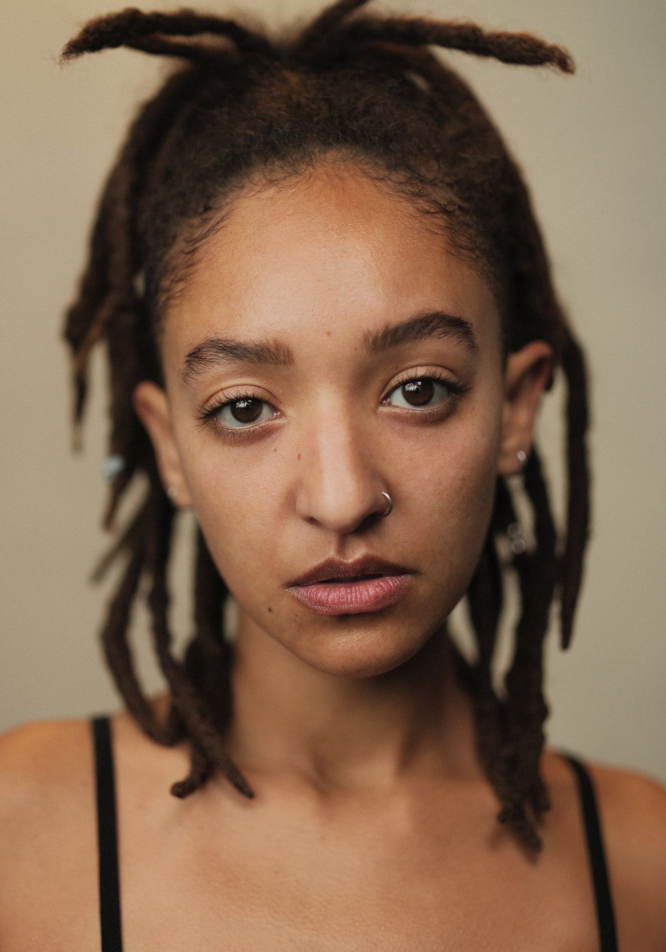 A neck up headshot on a beige background. They are about 30 with medium length dark brown hair in dreadlocks that are tied half up. They have brown eyes and a left silver nose ring. They are wearing a black camisole.
