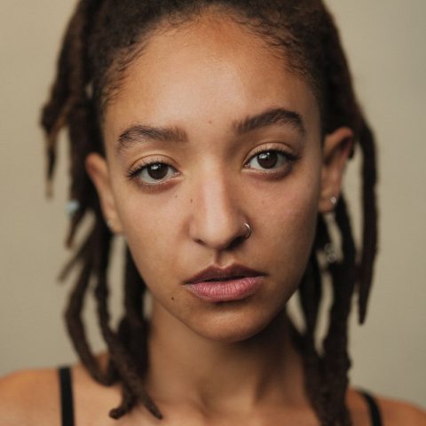 A neck up headshot on a beige background. They are about 30 with medium length dark brown hair tied half up. They have brown eyes and a left silver nose ring. They are wearing a black camisole.
