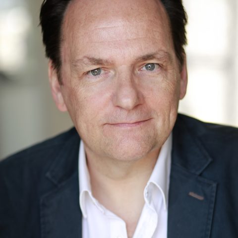 A chest up headshot on a blurred background. They are middle aged with short dark brown hair and green eyes. They are wearing a white collared shirt under a dark blue suit jacket.