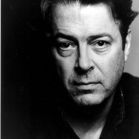 A black and white neck up headshot on a dark background. They are middle aged with short dark hair and dark eyes. They are wearing a black shirt.