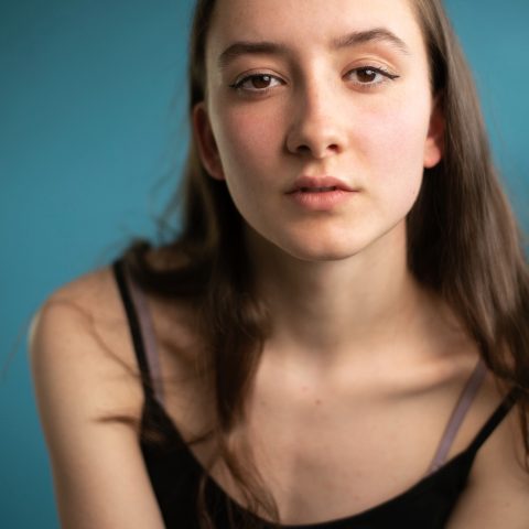 A chest up headshot on a teal background. They are about 25 with long straight brown hair and brown eyes. They are wearing a black camisole.