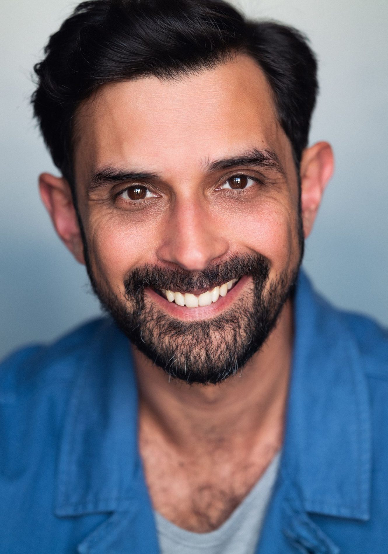 A neck up headshot on a light blue background. They are middle aged with short black hair, brown eyes, and a short beard and moustache. They are wearing a grey t-shirt under a blue collared shirt.