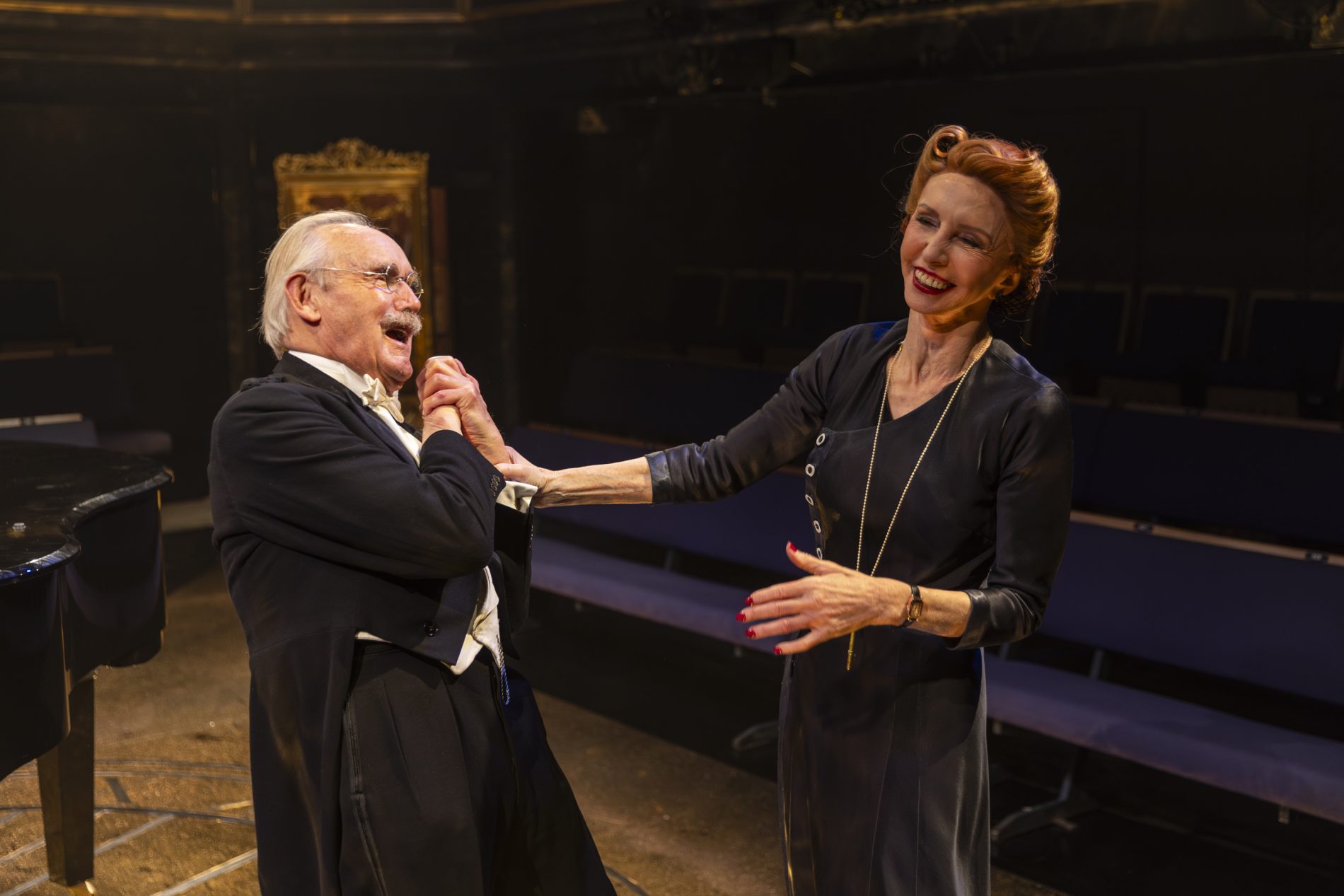 Jane Asher and Clive Francis laugh with each other. On the left is Clive Francis with short grey hair and a moustache. He is wearing a white collared shirt, white bowtie, white vest, dark trousers, black suit jacket, and round wire framed glasses. He is grasping both hands together in front of his chest. On the right is Jane Asher, with her auburn hair pulled into a neat updo. She is wearing a black dress, earrings, watch, and a long gold chain with a key. Her right hand is fully extended and holding Clive Francis' wrist.