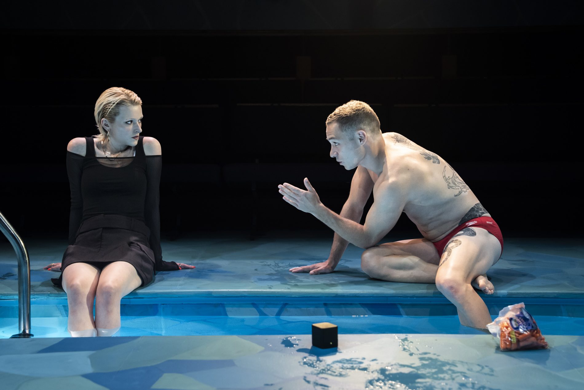 Two actors are on stage sitting at the edge of a pool. On the right an actor is seated with their feet in the pool and hands back behind them on the ground. They are about 30 with short slicked back blonde hair. They are wearing a black top and black skirt. They are looking at the other actor on the right with a serious expression. The other actor is about 30 with one leg in the pool. He has short brown hair, a large dragon back tattoo, and is wearing a red speedo. He is talking to the other actor; his hand pointing out towards them, expression serious. On the stage there is also a small black box and a bag of carrots.
