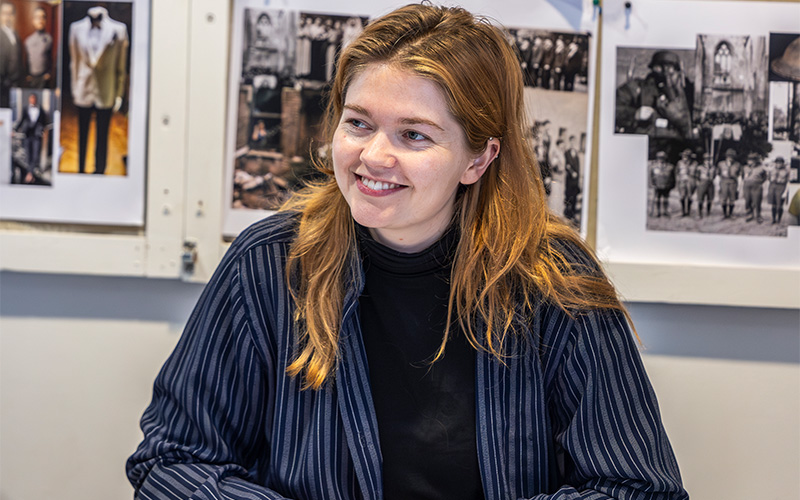 A close up shot of the assistant director looking and smiling at something off camera. They are about 25 with medium length auburn hair and wearing a black top under a navy blue striped collared shirt.