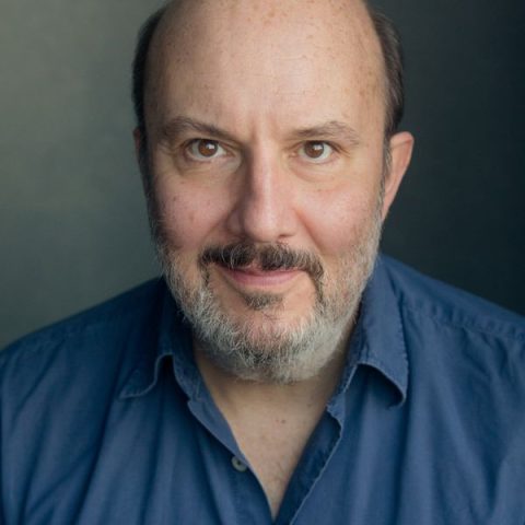 A chest up headshot on a dark grey background. They are middle aged with short brown hair, a grey and brown short beard and moustache, and brown eyes. They are wearing a blue collared shirt.