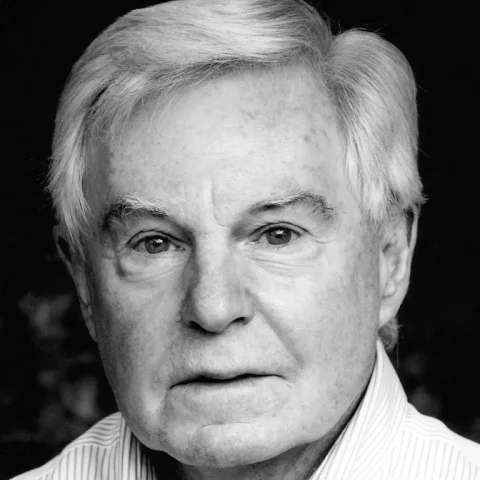 A black and white neck up headshot on a dark background. They are older with white hair and dark eyes. They are wearing a striped collared shirt.