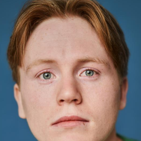 A headshot of an actor with short red hair and green eyes. They are wearing a green shirt and a neutral expression.