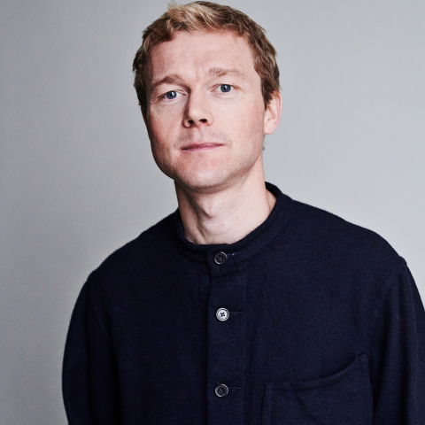 A headshot of an actor with short dirty blond hair who is wearing a navy sweater. they are wearing a neutral expression.