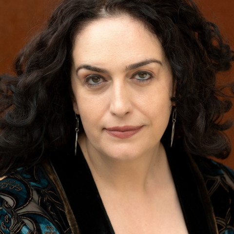 A headshot of an actor with long curly black hair. They are wearing a black blouse and have a mysterious expression on their face.