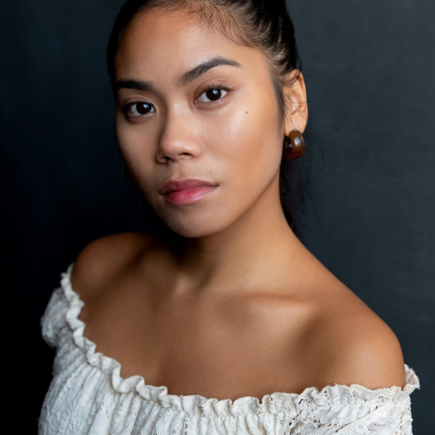 A headshot of an actor with black slicked black hair. They are wearing a white blouse and a neutral expression.