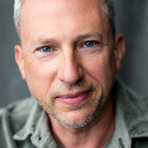 An actor with short grey hair, stubble, and blue eyes smiles into the camera. They are wearing a green collard shirt.