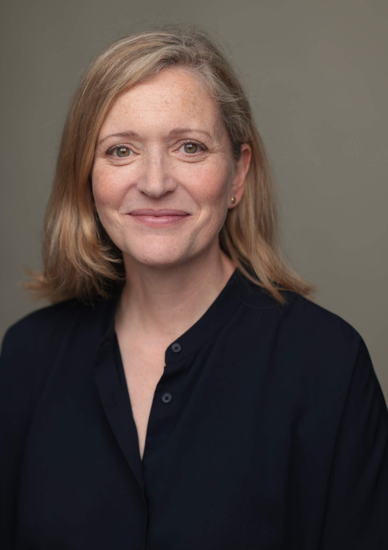 An actor with shoulder-length blonde hair smiles toward the camera against a medium grey background. They are wearing a black buttoned top, and are seen mostly from the shoulders-up.