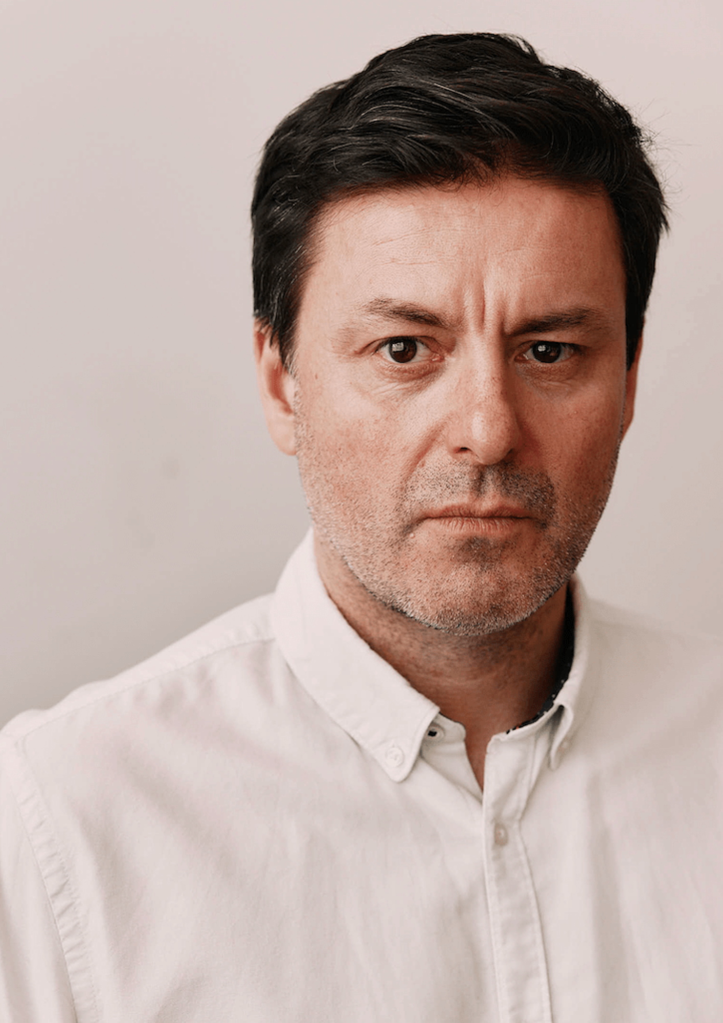 An actor with short brown hair has a neutral expression against a white background. They have a slight beard and are wearing a white buttoned shirt. They can be seen from the shoulders-up.