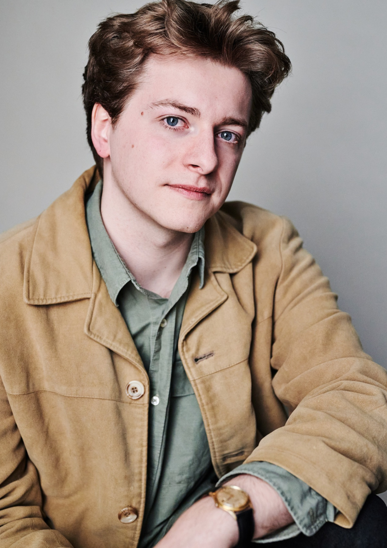 An actor with short brown hair gazes toward the camera with a neutral expression against a light grey background. They are wearing a green buttoned shirt under a light brown jacket. They have one arm resting casually on their knee, and can be seen mostly from the waist-up.