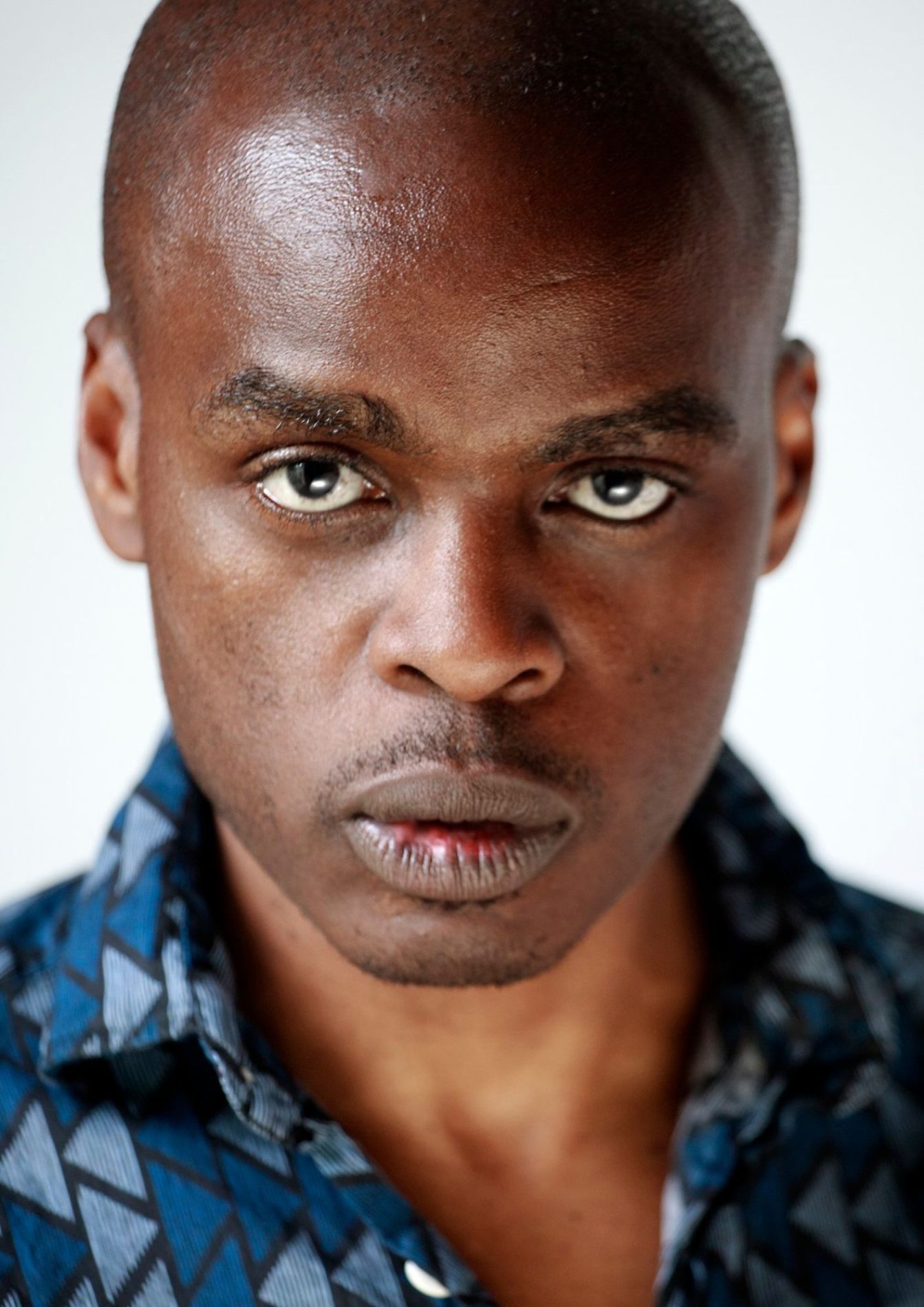 An actor with a buzz cut has an intense facial expression. They are wearing a blue shirt with multi-coloured triangles.