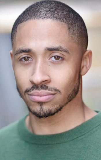 A headshot of an actor with short black hair, short beard, and brown eyes is wearing a green shirt and a serious expression.